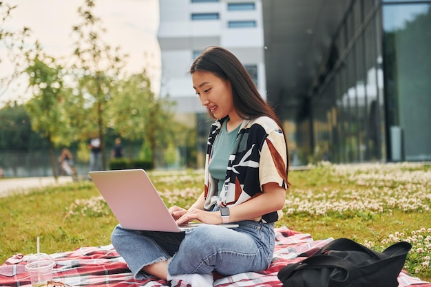Сидит с ноутбуком Молодая азиатская женщина находится на улице в дневное время
