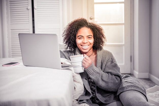 Sitting against laptop. Smiling beaming woman having cozy morning with cup of tea while working with laptop