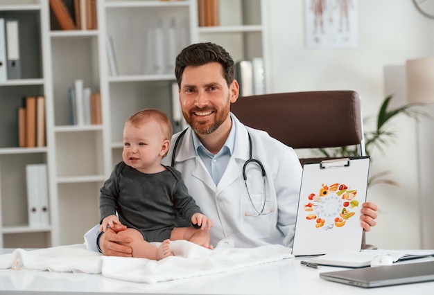 写真 座って微笑んでいる小さな赤ちゃんの医者はキャビネットで働いています