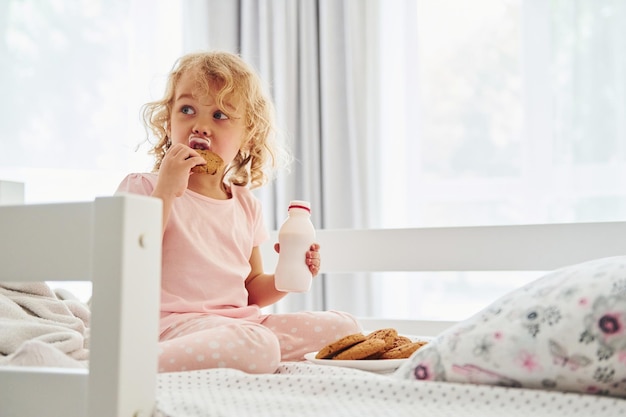 Sitting on the bed eating cookies and drinking milk Cute little girl in casual clothes is indoors at home at daytime