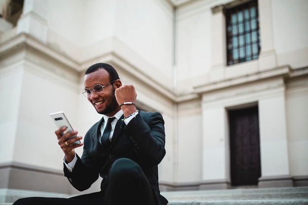 Sitting black businessman enjoys happiness for victory. Good news via cell phone.