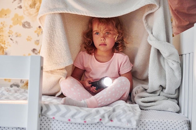 Sitting under blanket with flashlight Cute little girl in casual clothes is indoors at home at daytime