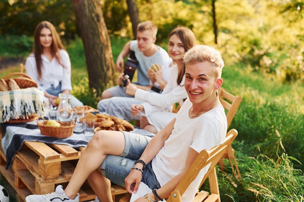 Sitting by picnic table Group of young people have vacation outdoors in the forest Conception of weekend and friendship