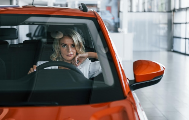 Sitting in the car Woman in formal clothes is indoors in the autosalon