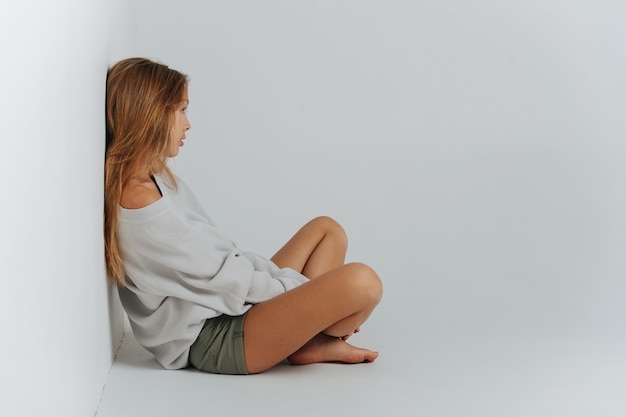 Sitting cross-legged on the floor teenage girl leaning her back against the wall. She's wearing shorts and warm longsleeve covering her body down to her thighs with bare legs showing under.