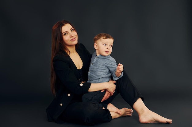 床に座っているスタイリッシュな黒い服を着た母親は、スタジオで彼女の幼い息子と一緒にいます