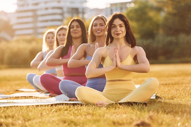 Photo sitting on the mat and meditating group of women have fitness outdoors on the field together