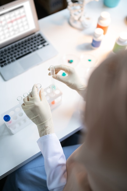 Sitting near laptop. Top view of chemist wearing gloves sitting near laptop and placing test tubes