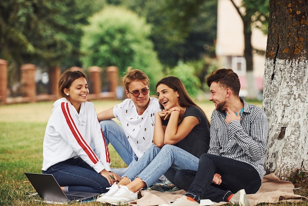 Sitting near the tree. Group of young students in casual clothes on green grass at daytime.