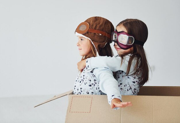 Sitting in the paper box Two cute little girls indoors at home together With retro pilot glasses and hat