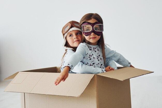 Sitting in the paper box Two cute little girls indoors at home together With retro pilot glasses and hat