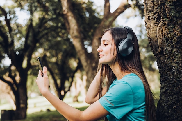 Sitting smiling teenage girl using headphones and listening music on her smartphone in the forest