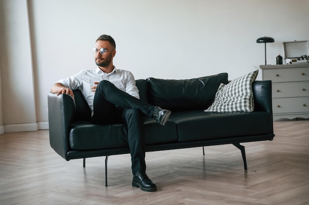 Photo sitting on the sofa elegant man in business clothes is indoors with glass of water