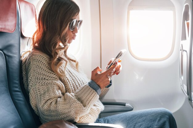 Сидя с телефоном в руках Молодая женщина-пассажир в повседневной одежде находится в самолете