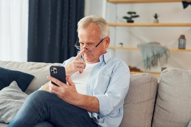 スマートフォンを手に座っている素敵な服を着た年配の男性が家にいる