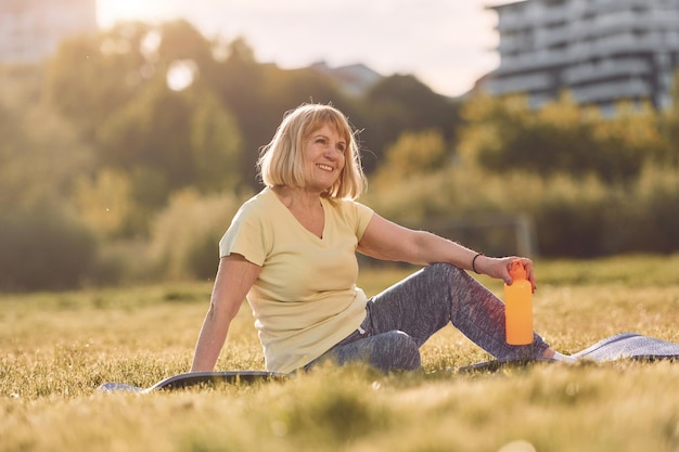 Sitting on yoga mat and holdng a bottle senior woman having\
nice weekend outdoors on the field at sunny day