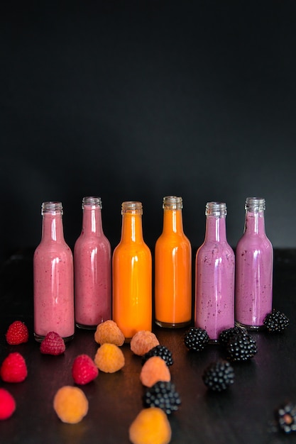 Six bottles with smoothies and raspberry, (red, yellow, blackberry) on black . Milk shake in glass jar with berries. diet or vegan food , fresh vitamin.