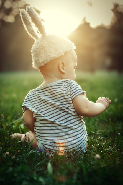 a six-month-old baby sits on the grass in the sunlight in a bunny hat with beautiful ears
