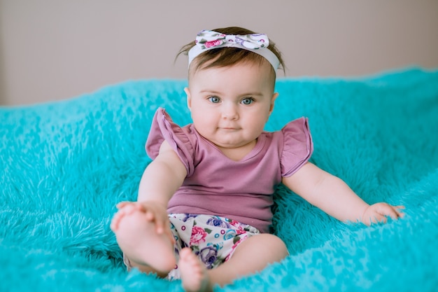 Six months old baby with creative ribbon on her head sitting on bed and looking at camera