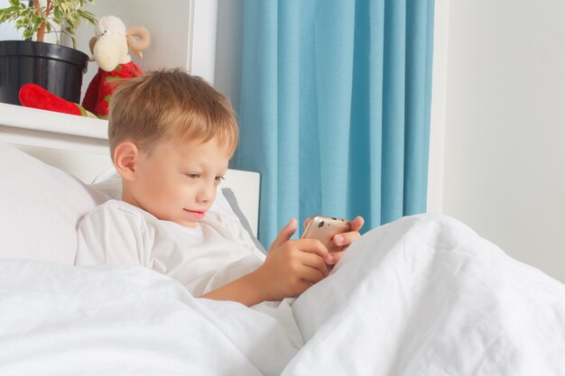 A six-year-old boy is resting lying in bed and holding a smartphone in his hands. A child with a gadget at home. 