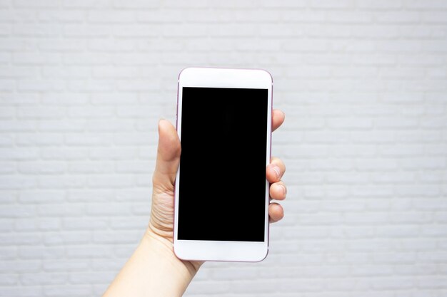 Sjabloon voor het ontwerp witte telefoon met zwart scherm in vrouwelijke hand op witte bakstenen muur