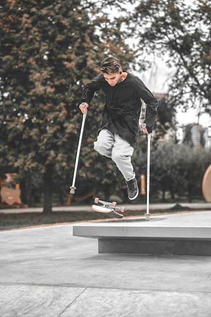 Skateboarden, inclusiviteit. Gerichte dappere man in zwart sweatshirt met handicap op krukken die 's middags op skateboard van springplank naar parauk springt