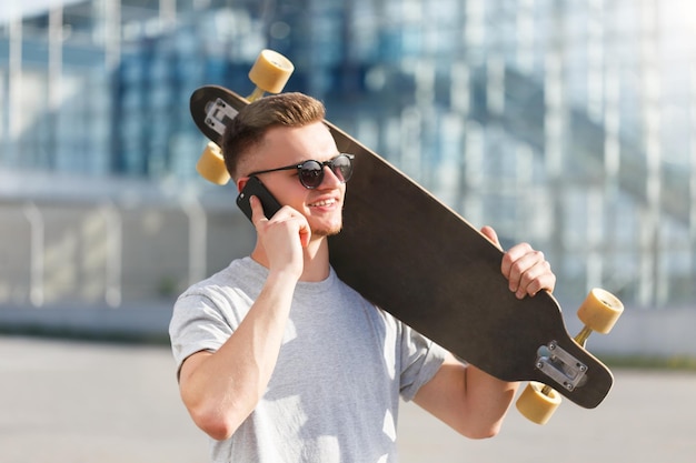 Skateboarder gekleed in grijze tshirt en zonnebril met longboard en praten door smartphone voor modern gebouw