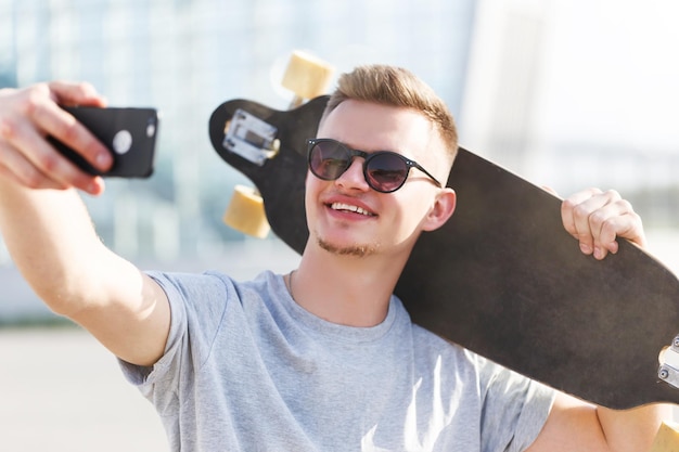 Skateboarder gekleed in grijze tshirt en zonnebril selfie maken op smartphone met longboard op schouder