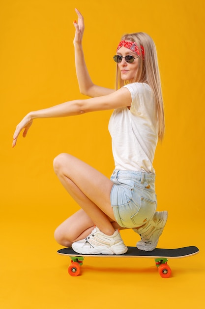 Skateborder girl in shorts and T-shirt sitting on board over yellow background