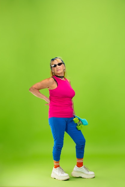 Skater girl. Senior woman in ultra trendy attire isolated on bright green background. Looks stylish and fashionable, forever young. Caucasian mature woman in sunglasses, bright attire and sneakers.