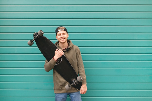 Skater in casual kleding staat met een longboard in zijn hand tegen de achtergrond van een turquoise heldere muur