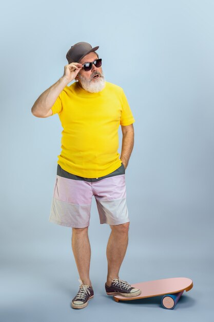 Skater man. Portrait of senior hipster man in eyewear isolated on light blue studio background. Tech and joyful elderly lifestyle concept. Trendy colors, forever youth. Copyspace for your ad.