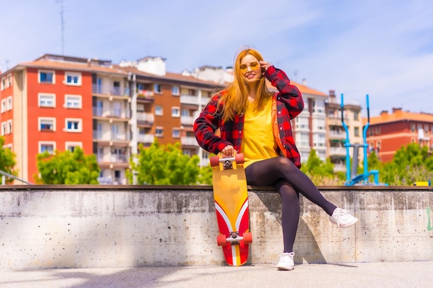 Skater vrouw in een geel t-shirt rood geruit hemd en zonnebril zittend met skateboard op een bankje in de stad glimlachend kijkend naar de camera