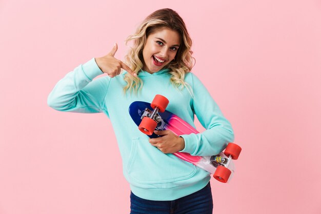 Skater woman smiling and holding skateboard, isolated over pink wall