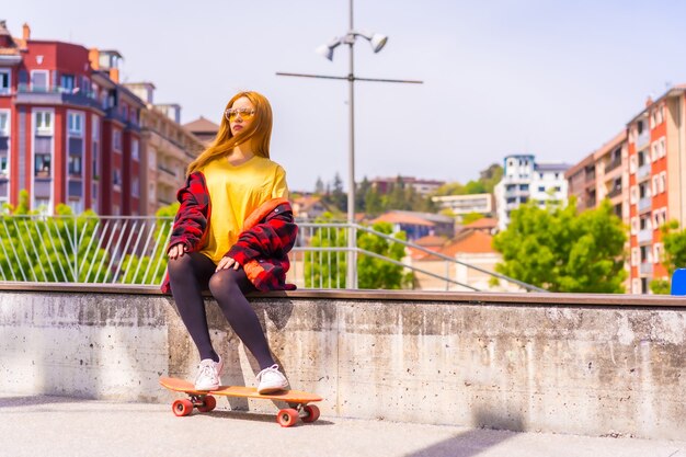 Skater donna in una t-shirt gialla, camicia a quadri rossa e occhiali da sole, seduta con lo skateboard su una panchina in città