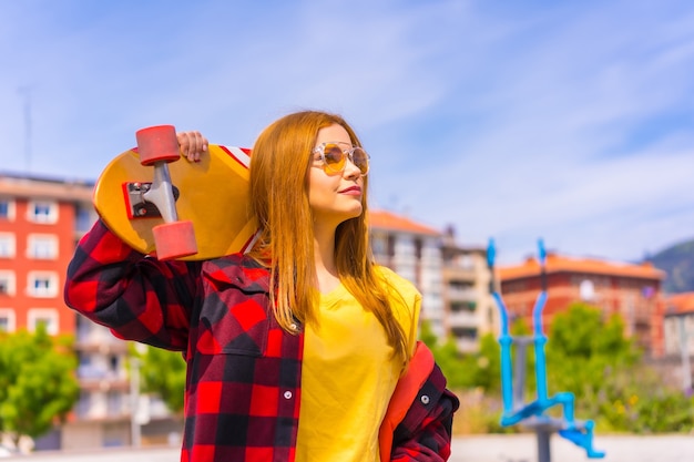 Skatervrouw in een geel t-shirt, rood geruit overhemd en zonnebril, peinzend naar rechts kijkend