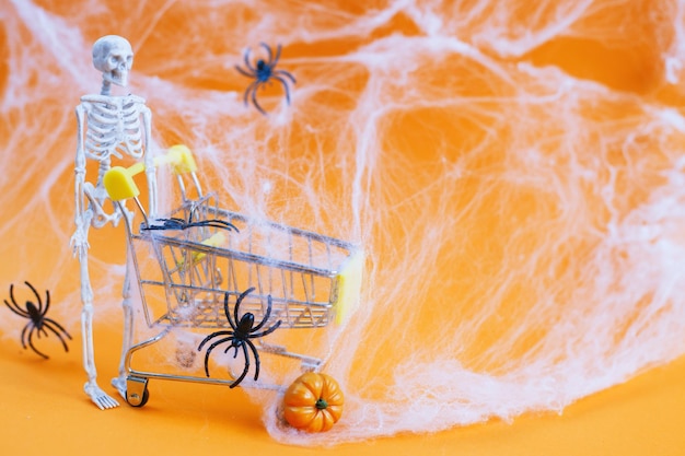 Skelet met een pompoen in de buurt van de trolleyspinnenwebben en spinnen op een oranje achtergrond Halloween-winkelconcept