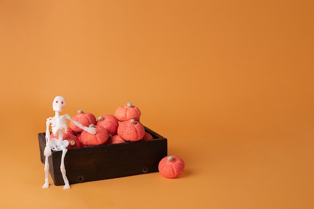 Skelet zittend op houten kist met marshmallow pompoenen op oranje achtergrond, halloween concept.