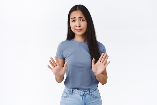 Foto donna asiatica scettica, arrogante e non divertita, alzando le braccia in stop, movimento di rifiuto, sorrisetto e accigliata offerta di antipatia giudicante, disinteressato al prodotto, muro bianco in piedi