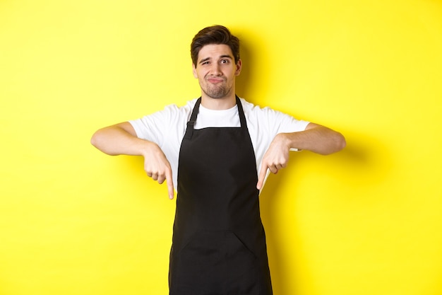 Skeptical barista in black apron pointing fingers down at bad product, looking displeased and unamused, standing over yellow wall