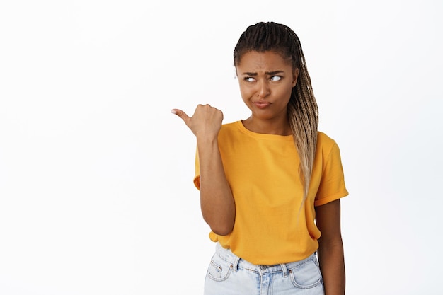 Skeptical Black girl with braids pointing and looking left smirking with displeased face expression white background