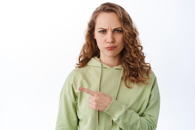 Skeptical blond girl frowning displeased pointing left at promotional text and look judgemental being disappointed in something standing over white background