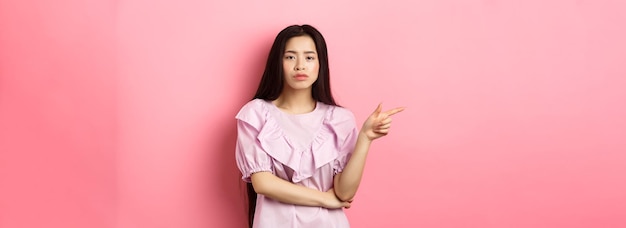 Skeptical chinese girl pointing finger left looking unamused and serious showing offer standing in d