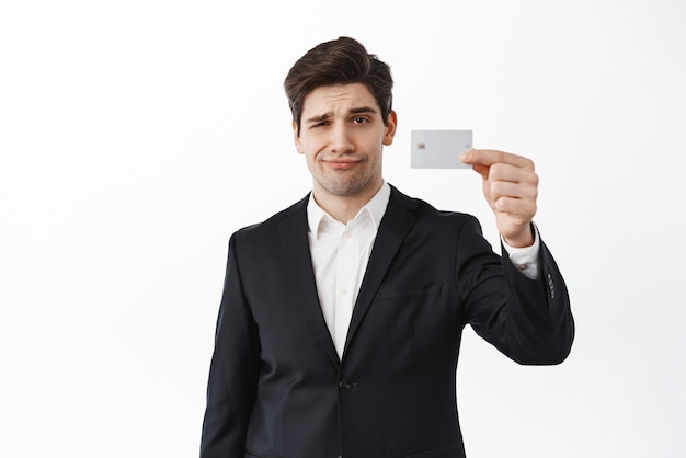 Skeptical displeased male entrepreneur business man shows credit card and frowning upset dislike bank standing over white background