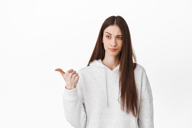 Skeptical and doubtful woman thinking looking hesitant at something strange pointing left and smirk with doubt being unsure standing over white background