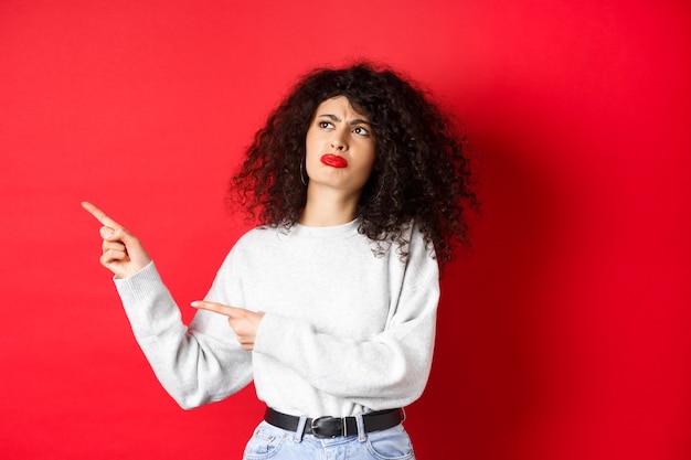 Skeptical frowning girl with curly hair, pointing and looking left hesitant, standing upset on red background.