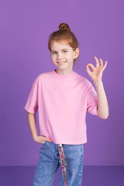 A skeptical little girl dressed in a pink Tshirt and jeans stands against the purple background of the studio holding her hands on her belt