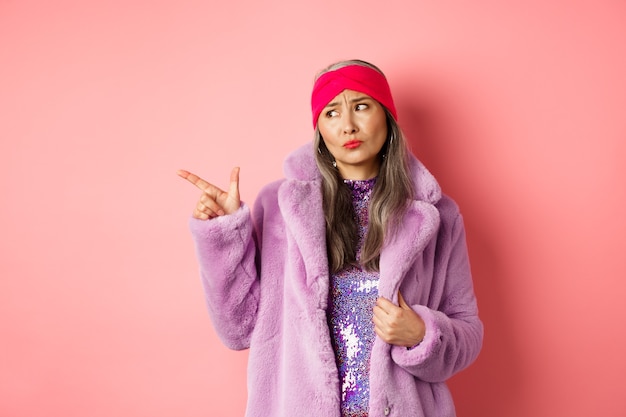 Skeptical middle-aged asian woman in purple fur coat looking and pointing finger left with displeased, sad expression, standing on pink.