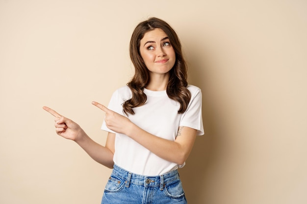 Skeptical modern girl, smirking, looking uncomfortable and pointing fingers left, showing advertisement, standing over beige background.