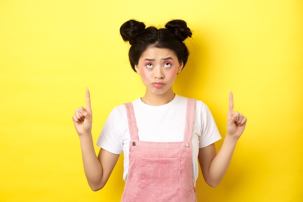 Skeptical teenage asian girl with glam pink makeup, pointing and looking up unamused, standing reluctant on yellow.